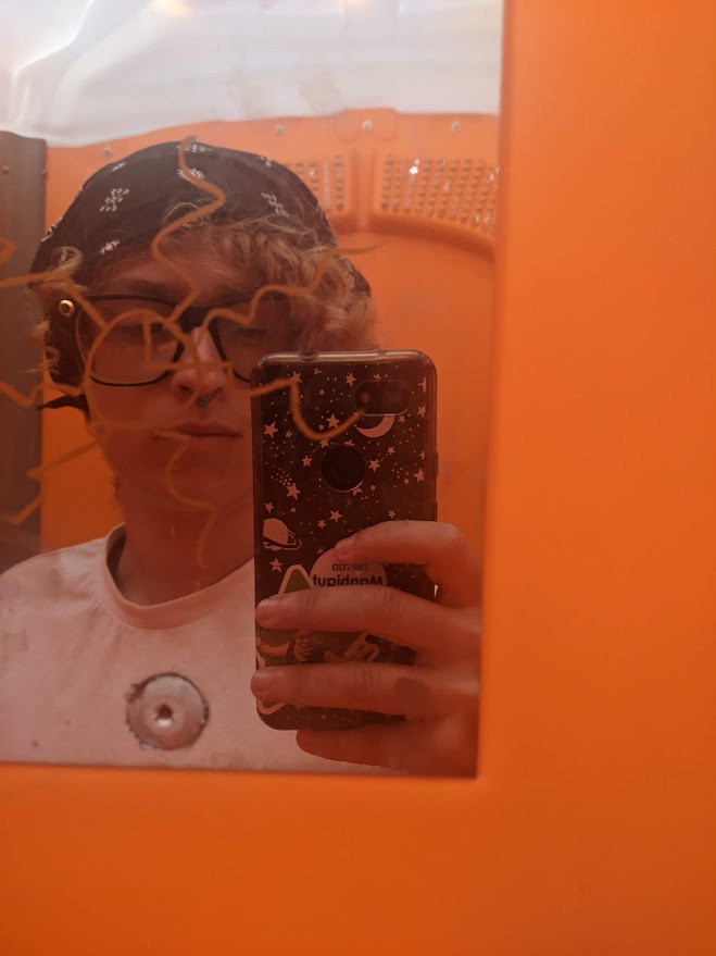 A white transman with pale pink hair wrapped in a black bandana, black glasses, and a white t-shirt takes a mirror selfie on his phone, which is in a galaxy themed case and has a Shrek sticker on it. The mirror has an orange sun drawn on it, partially obscuring his face, and the background is the same shade of bright orange.